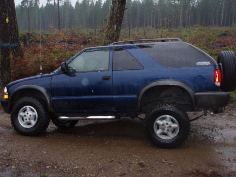 1995 Ford Bronco - A blue SUV, likely a Chevrolet Blazer from the early 2000s, is parked on a muddy road surrounded by trees.