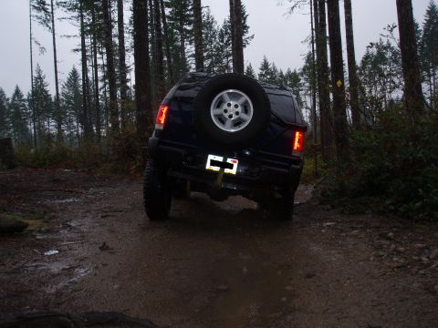 1995 Ford Bronco - A dark blue SUV navigates a muddy path through a dense forest, its spare tire mounted prominently on the rear.