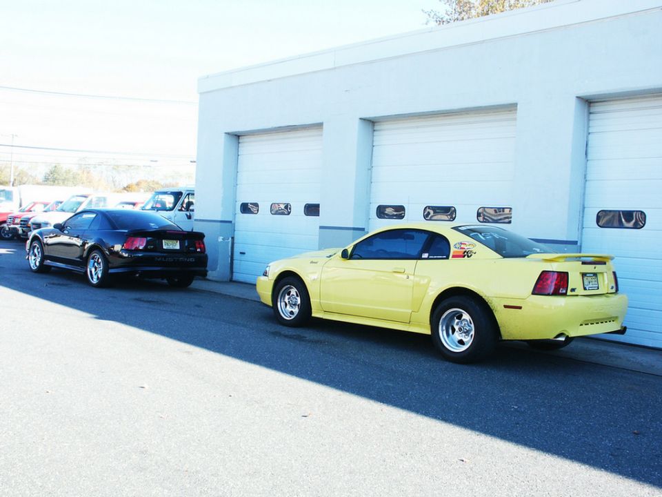 Yellow Ford Mustang parked next to a black one in front of garage doors. The yellow one has racing decals on the side