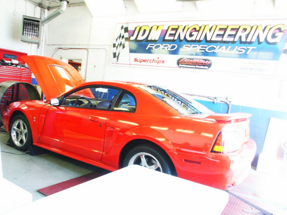 Red Ford Mustang coupe on a dynamometer with hood open. JDM Engineering sign in background