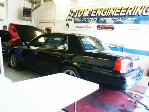 2001 Mercury Grand Marquis - A black sedan is positioned on a lift inside a garage, with a banner for JDM Engineering visible on the wall.