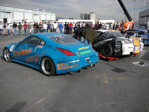 1998 Ford Crown Victoria - blue widebody drift car with sponsor decals sits among other race cars in an outdoor paddock as a crowd gathers.