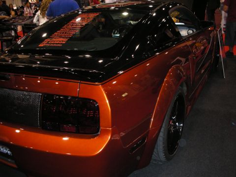 1998 Ford Crown Victoria - rear quarter of a copper-orange widebody Mustang with a black roof and smoked taillights at an indoor show.