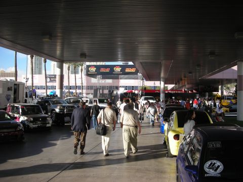 1998 Ford Crown Victoria - People walk beneath a covered outdoor concourse flanked by parked show vehicles, with palm trees and banners visible in the bright backgrou…