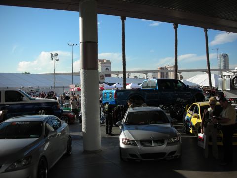 1998 Ford Crown Victoria - Row of show cars and a lifted blue crew-cab pickup parked under a covered venue, with event tents and a clear blue sky in the background.