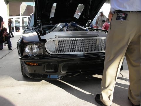 1998 Ford Crown Victoria - Low front view of a black custom car with a chrome mesh grille and round headlight, hood raised to show the engine, with onlookers standing…