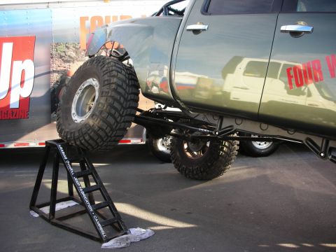 1998 Ford Crown Victoria - Rear of a lifted pickup raised on a display ramp next to a Four Wheeler magazine trailer, showing a large mud tire and long-travel suspensi…