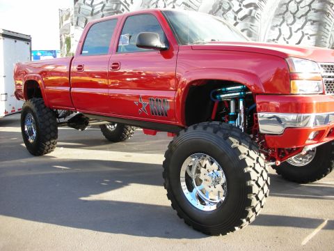 1998 Ford Crown Victoria - Heavily lifted red Chevrolet crew-cab pickup with a tube front end, blue coilover shocks, chrome wheels, and oversized mud tires on display.