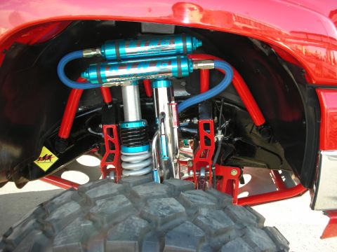 1998 Ford Crown Victoria - Close-up of a red truck's long-travel front suspension showing blue coilover and bypass shocks, red control arms, and mud tires.