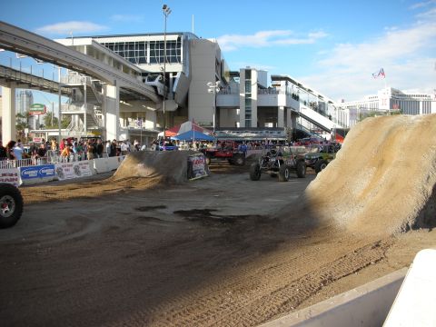 1998 Ford Crown Victoria - Outdoor dirt off-road demo course with tube-frame buggies on a packed track, spectators along the rail, and a city skyline in the backgroun…