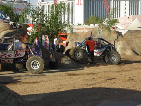 1998 Ford Crown Victoria - Two tube-frame rock buggies parked on a dirt off-road course beside potted palms, with hotel buildings and event banners behind.