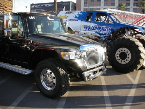 1998 Ford Crown Victoria - Black lifted military-style truck marked '460' parked beside a blue monster truck on huge tires at an outdoor display with Nitto and Pro Co…