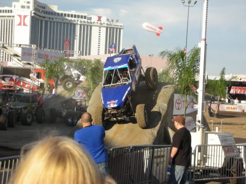 1998 Ford Crown Victoria - Blue tube-frame rock buggy launching off a dirt obstacle on an outdoor course as spectators watch, with casino hotels in the background.