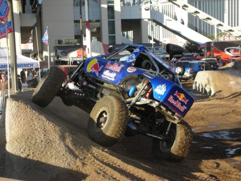 1998 Ford Crown Victoria - Blue Red Bull-liveried tube-frame rock buggy tilted on a dirt obstacle during an outdoor demo, with vendor tents and banners around the cou…