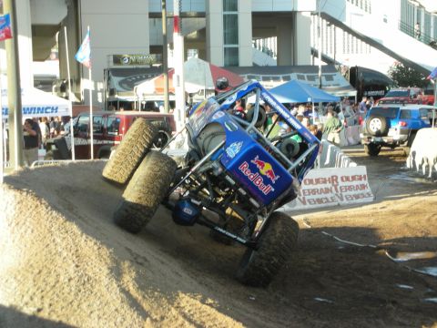 1998 Ford Crown Victoria - Blue Red Bull tube-frame rock buggy lifting a front wheel while crawling a dirt obstacle, with 'Tough Terrain Vehicle' signage and tents be…