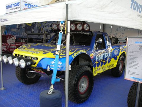 1998 Ford Crown Victoria - Blue and yellow Fabtech/Toyo Tires off-road race truck with light bars and large tires, displayed on blue flooring under a branded tent.
