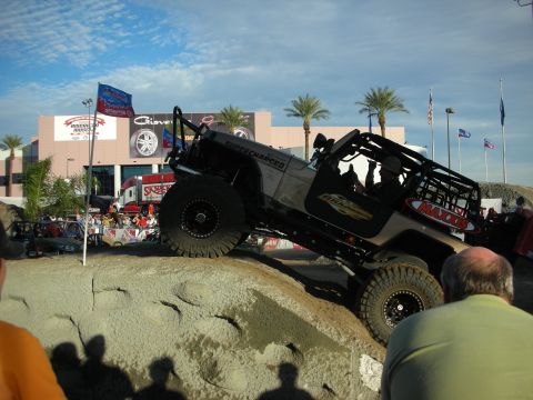 1998 Ford Crown Victoria - silver supercharged Jeep Wrangler crawls over a sculpted dirt mound on Maxxis tires while spectators watch at an outdoor show.