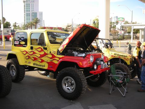 1998 Ford Crown Victoria - lifted yellow Jeep Wrangler with red flame graphics and BFGoodrich tires sits with its hood open at an outdoor display.