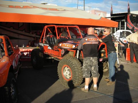 1998 Ford Crown Victoria - orange Maxxis-branded tube-chassis off-road race buggy sits under a canopy as men in pit gear talk beside it.