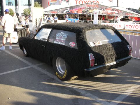 1998 Ford Crown Victoria - black wagon entirely covered in shaggy fur sits in a parking lot with Toyota and SEMA-style banners behind it.