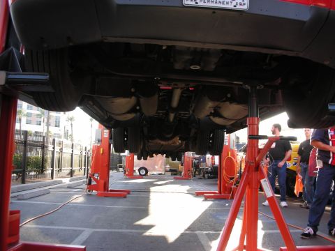 1998 Ford Crown Victoria - vehicle is hoisted high on red mobile column lifts, exposing its driveline and exhaust, with people gathered nearby outdoors.