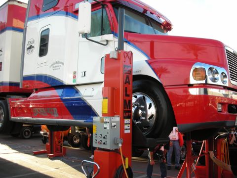 1998 Ford Crown Victoria - cab of a red, white and blue Freightliner-style semi tractor sits hoisted on red ALM column lifts at an outdoor event.