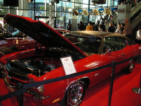 1998 Ford Crown Victoria - red Chevelle SS muscle car with its hood up and black vinyl roof sits roped off on a red carpet at an indoor show.