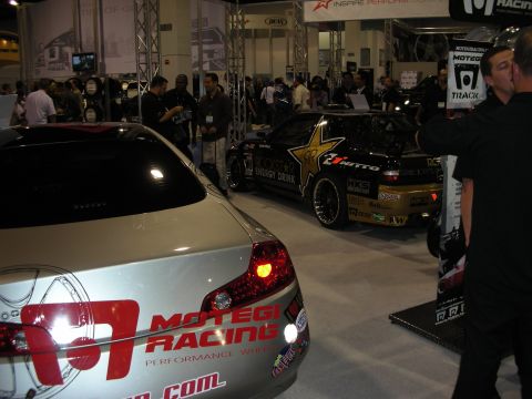 1998 Ford Crown Victoria - silver Motegi Racing sedan sits in the foreground while a gold Rockstar Energy drift car shows behind it at an indoor booth.