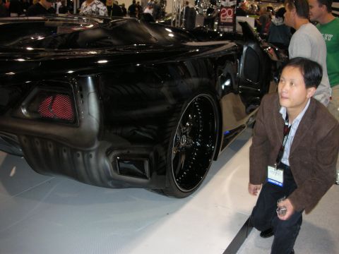 1998 Ford Crown Victoria - black custom concept car with large black wheels sits on a white display floor while a man in a brown jacket admires it.