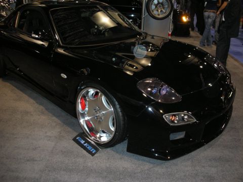 1998 Ford Crown Victoria - glossy black Mazda RX-7 with polished multi-spoke wheels sits on a Falken-branded stand at an indoor show.