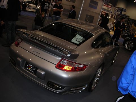 1998 Ford Crown Victoria - silver Porsche 911 Turbo with a deployed rear wing sits on a dark carpet at an indoor show as people browse behind it.