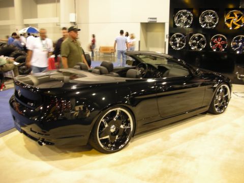 1998 Ford Crown Victoria - black two-door convertible on large chrome wheels parked before a wall of display wheels with onlookers nearby.