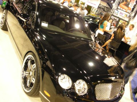 1998 Ford Crown Victoria - black Bentley sedan with chrome mesh grille and chrome wheels on display indoors as people sit at a booth nearby.
