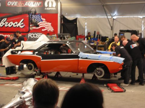 1998 Ford Crown Victoria - orange and black muscle car body on jack stands being worked on by a crew at an Overhaulin tent with sponsor banners.