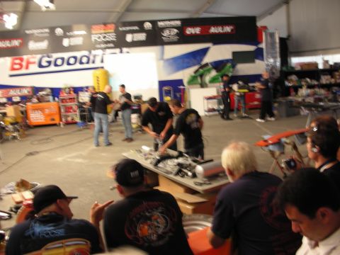 1998 Ford Crown Victoria - crowd watches builders work at tables inside an Overhaulin tent with BFGoodrich and sponsor banners overhead.