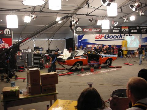 1998 Ford Crown Victoria - orange and black muscle car body on stands under stage lights and BFGoodrich banners inside an Overhaulin event tent.