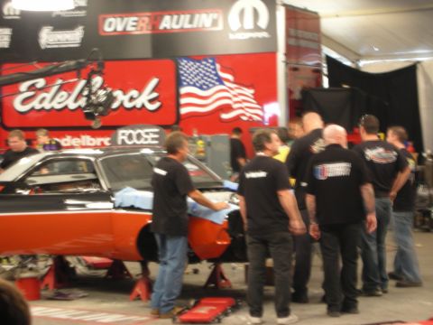 1998 Ford Crown Victoria - black-shirted crew works on an orange and black muscle car body before Edelbrock and Overhaulin banners and a US flag.