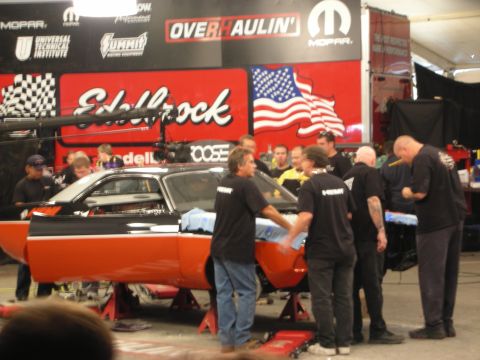 1998 Ford Crown Victoria - crew in black shirts gathers around an orange and black muscle car body before Edelbrock and Mopar Overhaulin banners.