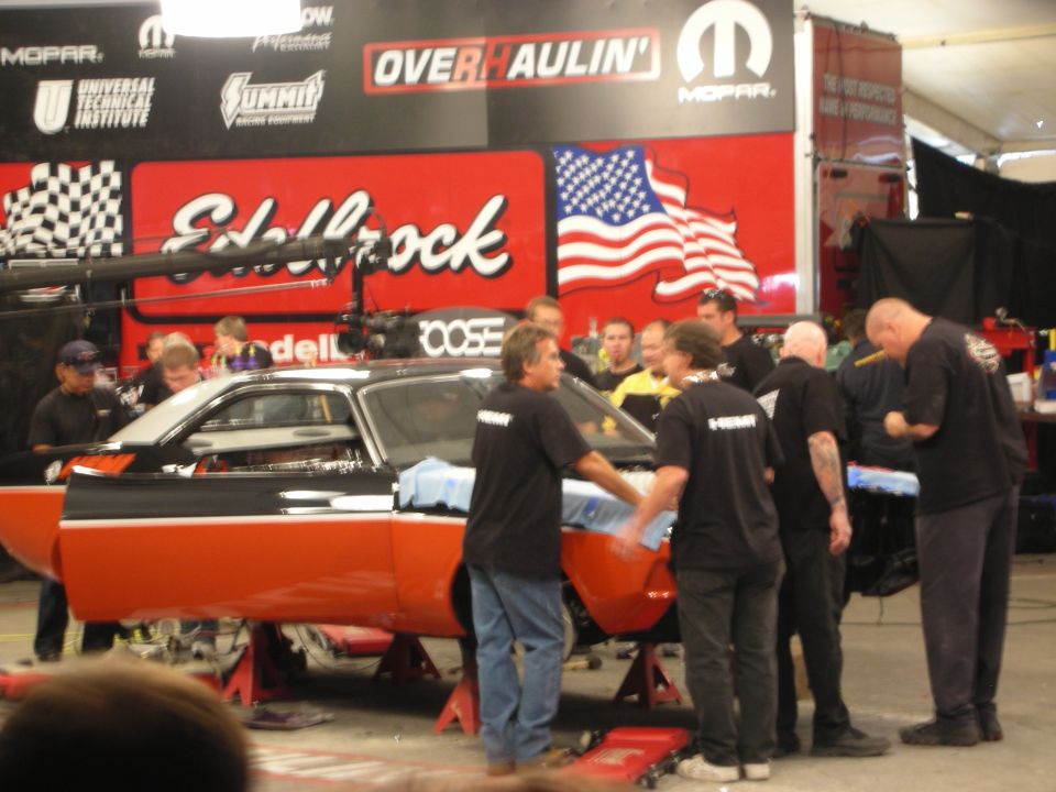 Crew gathered around orange muscle car at Edelbrock Overhaulin tent