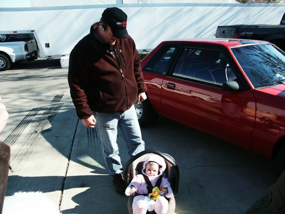 Red coupe with racing roll cage visible through the windows, parked next to a man and a baby in a car seat
