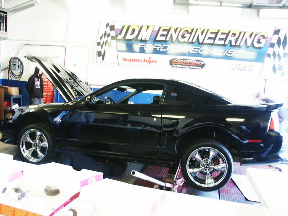 Black Ford Mustang coupe on a lift, hood open, chrome wheels, in a garage with JDM Engineering banner