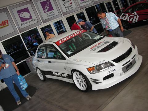 1998 Ford Crown Victoria - white Mitsubishi Lancer Evolution sedan with white wheels and HKS, Zeal, and sponsor decals on display outside a show hall.
