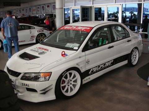 1998 Ford Crown Victoria - white Mitsubishi Lancer Evolution with sponsor decals, white wheels and a roll cage sits on display at an indoor automotive show.