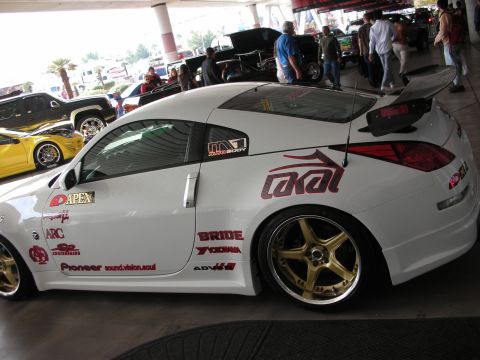 1998 Ford Crown Victoria - white Nissan 350Z coupe covered in sponsor decals shows off gold wheels and a tall rear wing under a covered show area.