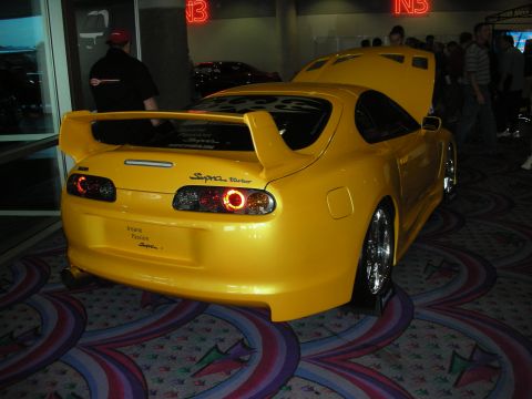 1998 Ford Crown Victoria - yellow Toyota Supra Turbo with a rear spoiler and ringed taillights sits on patterned carpet at an indoor venue.