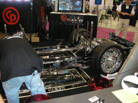 1998 Ford Crown Victoria - heavily chromed custom car chassis with hydraulic rams and large wheels sits on a black stage at a trade show booth.