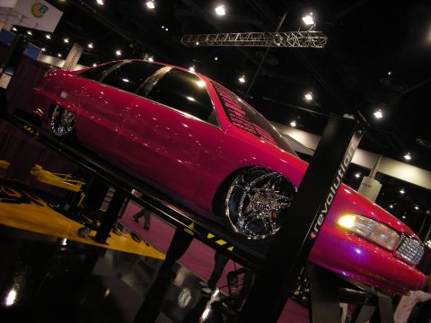 1998 Ford Crown Victoria - glossy pink custom sedan with chrome spinner wheels sits raised on a display lift under spotlights at an indoor show.
