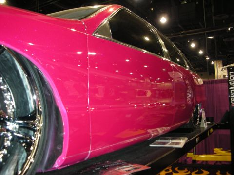 1998 Ford Crown Victoria - close-up of a glossy pink custom car shows its fender and chrome wheel beside a Revolution display banner indoors.