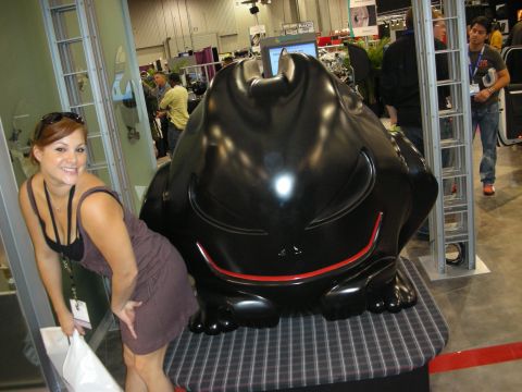 1998 Ford Crown Victoria - smiling woman in a gray dress poses next to a large glossy black cartoon-cat-shaped subwoofer enclosure at a trade show.