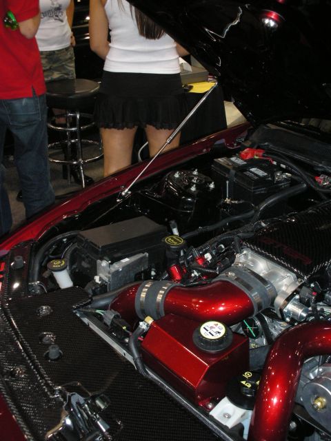 1998 Ford Crown Victoria - open engine bay with candy-red painted components and braided lines is shown as people stand beside the car at a show.